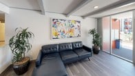 The El Cortez leasing office is a thoughtfully designed single-floor space that blends modern aesthetics with a welcoming, gallery-like atmosphere. The open floor plan is anchored by wood-look tile flooring that runs throughout, complemented by crisp white walls and distinctive exposed beam ceilings that add architectural character.

The reception area features a clean, white front desk accompanied by sleek bar-height seating and a chrome open-shelf display unit stocked with informational materials. Colorful circular artworks punctuate the walls, contributing to the space's vibrant, contemporary personality. A wall-mounted flat-screen display and a warm-toned credenza with wood-grain cabinetry create a multimedia presentation area ideal for showcasing the property.

A comfortable lounge seating area includes a tufted navy leather sectional sofa set beneath a large-scale abstract painting, flanked by tall indoor plants that bring a natural element into the space. Floor-to-ceiling glass sliding doors branded with the El Cortez logo separate an inner office area, providing both transparency and privacy. Large glass entry doors along the front facade allow abundant natural light to fill the interior, creating an inviting and energetic environment throughout.