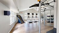 Located at 100 Antioch Pike in Nashville, TN, this fitness center is a thoughtfully designed, single-floor facility offering a comprehensive workout environment. The space is defined by its striking vaulted ceilings with exposed beams, which lend an airy, open feel throughout. Warm-toned hardwood-style flooring runs the length of the room, complementing the bright, neutral walls and creating a clean, modern aesthetic.

Natural light floods the interior through a series of large arched windows positioned along the walls, reducing the need for artificial lighting and giving the space an inviting ambiance. Pendant and ceiling-mounted lights, along with a ceiling fan, ensure the environment remains comfortable during all hours of use.

The cardio zone features a row of treadmills, a stair-climber machine, and additional aerobic equipment, each accompanied by wall-mounted flat-screen televisions for entertainment during workouts. The strength training area is equally well-appointed, with a Smith machine, cable systems, adjustable benches, barbells, weight plates, and a full rack of dumbbells. A functional training rig with pull-up bars and gymnastic rings rounds out the strength section.

Full-length mirrors line portions of the walls, enhancing the sense of space and supporting proper form during exercise. The layout is organized and spacious, allowing for fluid movement between equipment zones.