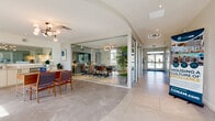 This single-story lobby and clubhouse in Rockledge, Florida, is designed with a contemporary aesthetic that blends functionality and comfort. The entrance greets visitors with soaring double-height ceilings, floor-to-ceiling windows, and large glass double doors that flood the space with natural light and provide views of the surrounding landscaping. Large-format tile flooring in a neutral stone finish runs throughout, creating a cohesive and polished look.

The lounge area features vibrant, modern furnishings, including a deep navy sofa, cognac leather armchairs, and a coffee table set against white walls adorned with coastal-themed artwork. Lush tropical plants are positioned throughout, enhancing the Florida-inspired ambiance. A dedicated seating nook with tall windows frames outdoor views of palms and greenery, offering a quiet retreat.

The game room includes a full-size pool table, arcade machines, and a wall-mounted television, all beneath a striking pendant light and soaring ceilings. Adjacent to this space is a glass-enclosed conference room, accessible via sliding glass doors and furnished with a large table and office chairs.

The bar area features a clean white countertop, light wood cabinetry, a seafoam-toned glass backsplash, a mounted television, and teal bar stools, creating a casual gathering spot. The overall ambiance is bright, modern, and welcoming throughout.