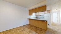 This single-floor unit offers a practical and straightforward layout designed for comfortable everyday living. The kitchen is a central feature, equipped with warm wood-toned lower cabinetry and lighter upper cabinets, gray laminate countertops, a double stainless steel sink, a white electric range, and a refrigerator. A peninsula-style breakfast bar separates the kitchen from the adjacent dining or living area, which is finished with earthy tan tile flooring. Upper cabinetry extends to the ceiling, maximizing storage capacity throughout the kitchen.

The living area is generously sized, finished with neutral beige carpet and a textured ceiling with flush-mount lighting. A full-width sliding glass door fitted with vertical blinds allows natural light into the space and provides exterior access. The open layout of this area offers flexibility for various furniture arrangements.

The bathroom is well-appointed with warm-toned tile flooring, a light wood vanity with a granite-style countertop, an integrated sink, a large mirror, and a full-size tub and shower combination with a curved curtain rod. The space is clean and functional, maintaining a consistent, understated aesthetic throughout the unit.