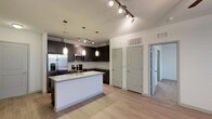 This single-floor, two-bedroom, two-bathroom residence offers a well-designed open-concept layout that seamlessly connects the kitchen and living areas. The kitchen serves as a central focal point, featuring dark espresso cabinetry that contrasts sharply with light countertops on the central island, which includes an undermount sink and provides ample workspace and casual seating potential. Stainless steel appliances, including a French door refrigerator, built-in microwave, and range, add a polished, contemporary feel. Track lighting runs along the ceiling, complemented by pendant lights suspended above the island, ensuring the kitchen is both well-lit and visually appealing. Light wood-tone flooring extends throughout the open living space, lending warmth and continuity to the interior. The layout includes multiple doors leading to private areas of the residence, including the two bedrooms, suggesting a thoughtful floor plan with clear separation between living and sleeping spaces. The overall aesthetic is modern and clean, with neutral tones and quality finishes creating a cohesive, move-in-ready environment.