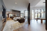 This upscale amenity space is thoughtfully designed to offer a variety of social and recreational experiences within a single, flowing floor plan. The design language is modern and refined, with warm wood-tone flooring running throughout and striking dark wood vertical slatted partitions that elegantly divide the space without fully enclosing it, maintaining an open and airy feel.

The main lounge area anchors the space with generous sectional and accent seating arranged over a textured area rug, bathed in natural light streaming through floor-to-ceiling windows that overlook the outdoors. Adjacent to the lounge is a dedicated game room featuring a full-sized billiard table beneath a sculptural branching chandelier, positioned in front of a wall-mounted television and a sleek linear fireplace set into large-format tile.

A separate gaming room enclosed by steel-framed glass partitions houses a round poker table with white accent chairs and its own wall-mounted screen. A well-appointed bar area offers a long quartz-topped counter with bar seating, a wood-paneled ceiling, and dual wall-mounted televisions — ideal for entertaining.

The space also includes a private dining area centered around a live-edge communal table illuminated by pendant lighting, alongside a catering kitchen. A quiet sitting nook with plush chairs and garden views rounds out this comprehensively appointed amenity floor.