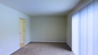 This two-floor property offers a straightforward and functional layout with clean, neutral finishes throughout. The pictured room features wall-to-wall carpeting in a warm beige tone, providing a comfortable and quiet underfoot feel. The walls are finished in a soft, light neutral color that contributes to a bright and airy atmosphere. White baseboards frame the perimeter of the room, adding a crisp, polished contrast to the muted wall tones. A set of vertical blinds on the right side of the room covers what appears to be a large window or sliding door, allowing adjustable control of natural light. A doorway on the left side of the room leads to an adjacent space with visible hardwood or hard-surface flooring, suggesting a transition between different functional areas of the home. A bi-fold door is partially visible, likely concealing a closet. The room's clean lines and open feel make it a versatile space adaptable to various uses. The two-floor configuration of the property provides additional separation of living areas, adding both functional space and a sense of scale to the overall home.