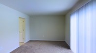 This two-floor property offers a straightforward and functional layout with clean, neutral finishes throughout. The pictured room features wall-to-wall carpeting in a warm beige tone, providing a comfortable and quiet underfoot feel. The walls are finished in a soft, light neutral color that contributes to a bright and airy atmosphere. White baseboards frame the perimeter of the room, adding a crisp, polished contrast to the muted wall tones. A set of vertical blinds on the right side of the room covers what appears to be a large window or sliding door, allowing adjustable control of natural light. A doorway on the left side of the room leads to an adjacent space with visible hardwood or hard-surface flooring, suggesting a transition between different functional areas of the home. A bi-fold door is partially visible, likely concealing a closet. The room's clean lines and open feel make it a versatile space adaptable to various uses. The two-floor configuration of the property provides additional separation of living areas, adding both functional space and a sense of scale to the overall home.
