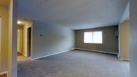 This single-floor space offers a clean, functional layout designed for comfortable everyday living. The main living area is generously sized, featuring wall-to-wall carpeting in a warm neutral tone that adds softness underfoot and contributes to the room's understated, welcoming atmosphere. The walls are painted in a light grey-beige palette, creating a calm and versatile backdrop suitable for a variety of interior styles. A textured ceiling adds a subtle layer of detail overhead. Natural light enters through a centrally positioned window, brightening the space and providing a connection to the outdoors. A baseboard heating system runs along the exterior wall beneath the window, ensuring efficient and consistent warmth throughout the colder months. A doorway on one side of the main room leads to additional areas of the unit, suggesting a practical and accessible floor plan. The overall ambiance is neutral and move-in ready, offering a blank canvas that accommodates a range of furnishing and decorating preferences. The space is straightforward in its design, prioritizing functionality and comfort in equal measure.
