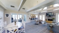 This single-floor space offers an open, airy layout highlighted by vaulted ceilings with exposed beams that add architectural character and a sense of spaciousness. The interior is bathed in natural light, courtesy of multiple sets of French doors and large windows lining the perimeter, creating a welcoming and open atmosphere throughout.

The space is divided into distinct functional zones that flow seamlessly into one another. A casual dining area near the entrance features a round white table with white cross-back chairs, grounded by a patterned blue and gray wave-motif carpet that extends throughout the interior. The lounge area is furnished with plush gray swivel chairs and a glass-top coffee table, offering a comfortable setting for relaxed conversation.

Toward the rear, a bar counter with tall gray barstools is tucked into a recessed nook illuminated by three pendant lights with warm gold fixtures, creating an inviting focal point. A separate glass-paneled door leads to an adjoining private room. The overall design aesthetic is coastal-inspired, combining soft neutrals, cool blues, and gold accents for a polished yet relaxed ambiance.