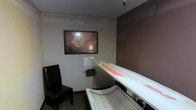 This single-floor tanning room offers a private, self-contained space designed for personal tanning sessions. The room features a full-size professional tanning bed as its centerpiece, equipped with high-output lamps for effective UV exposure. The interior is finished with neutral light grey and warm brown accent walls, creating a calm, understated ambiance. A framed botanical artwork hangs on the wall, adding a decorative touch to the otherwise functional space. A black cushioned chair is positioned in the corner, providing a place to sit before or after a session. The room is compact yet efficiently laid out, allowing easy access to the tanning bed from all sides. Subtle overhead lighting and a small mounted unit on the wall — likely a control panel or timer — contribute to the room's practical design. The flooring features a patterned tile, adding a modest visual element to the space. Overall, the room is straightforward and purpose-driven, providing a clean and private environment suited for individual tanning use.