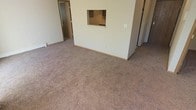 This single-floor residence offers a clean, functional layout designed with everyday comfort in mind. The living area is generously sized, featuring wall-to-wall carpeting in a warm, neutral tone that adds a cozy feel underfoot and ties the space together cohesively. The walls are finished in a soft off-white, creating a bright and airy atmosphere throughout.

A standout architectural detail is the pass-through window cut into the dividing wall between the living area and the kitchen, allowing for easy communication and a sense of openness between the two spaces. The kitchen area, visible through the pass-through, features light cabinetry and practical countertop space, making it both efficient and accessible.

A doorway off the living area leads to additional rooms, including what appears to be a bathroom with a frosted glass window, suggesting thoughtful privacy considerations in the design. A hallway visible along one side of the space hints at further rooms beyond.

Neutral baseboards frame the carpeted floors throughout, contributing to the overall clean and well-maintained aesthetic. The space benefits from a straightforward, open feel that lends itself well to a variety of furniture arrangements. Overall, this residence presents a practical and welcoming environment with a cohesive, understated design throughout.