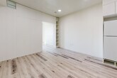 This thoughtfully designed single-floor unit offers a clean, modern aesthetic throughout, with light wood-look flooring and crisp white walls creating a bright, cohesive atmosphere across all spaces. The kitchen is efficiently laid out with white lower cabinetry, contrasting grey upper cabinets, a light stone-finish countertop, a stainless steel undermount sink, a black induction cooktop, a built-in Samsung microwave oven, and a ventilation hood — providing a fully functional cooking space despite the compact footprint. Recessed lighting illuminates the open corridor connecting the kitchen to the living and sleeping areas. The bedroom benefits from a large picture window that invites abundant natural light and frames outdoor views of the surrounding neighborhood, complemented by a wall-mounted HVAC unit for climate control. A versatile secondary room features a built-in open shelving unit, offering flexible storage or display space, and is separated from adjacent areas by a sliding panel door. The bathroom is accessed through a sleek door with a brushed metal bar handle and includes a soaking tub with surround, a wall-mounted toilet, and grab bars — all finished in a clean, neutral palette. Consistent wood-look flooring flows throughout, unifying the space.