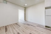 This thoughtfully designed single-floor unit offers a clean, modern aesthetic throughout, with light wood-look flooring and crisp white walls creating a bright, cohesive atmosphere across all spaces. The kitchen is efficiently laid out with white lower cabinetry, contrasting grey upper cabinets, a light stone-finish countertop, a stainless steel undermount sink, a black induction cooktop, a built-in Samsung microwave oven, and a ventilation hood — providing a fully functional cooking space despite the compact footprint. Recessed lighting illuminates the open corridor connecting the kitchen to the living and sleeping areas. The bedroom benefits from a large picture window that invites abundant natural light and frames outdoor views of the surrounding neighborhood, complemented by a wall-mounted HVAC unit for climate control. A versatile secondary room features a built-in open shelving unit, offering flexible storage or display space, and is separated from adjacent areas by a sliding panel door. The bathroom is accessed through a sleek door with a brushed metal bar handle and includes a soaking tub with surround, a wall-mounted toilet, and grab bars — all finished in a clean, neutral palette. Consistent wood-look flooring flows throughout, unifying the space.
