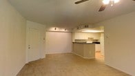 This single-floor residence features an open-concept design that seamlessly connects the main living area to the kitchen. The living space is finished with wall-to-wall beige carpeting, contributing to a neutral and understated aesthetic. A ceiling fan is installed overhead, providing practical air circulation throughout the room. Track lighting along the ceiling adds directed illumination to complement the overall lighting scheme.

The kitchen is separated from the living area by a raised peninsula counter with a dark countertop, offering a natural division between the two spaces while maintaining an open feel. White cabinetry lines the kitchen walls, providing ample storage, and is paired with white appliances including a range and refrigerator. The kitchen floor transitions to hard flooring, contrasting with the carpeted living area. A drop ceiling with fluorescent lighting brightens the kitchen workspace.

The walls throughout are painted in a soft, neutral tone, and the textured ceiling adds a subtle visual element consistent across the space. An entry door is visible along one wall, and HVAC vents are positioned to ensure climate control throughout the unit.
