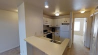 This single-floor space centers around a practical and well-organized kitchen designed for efficient use. The kitchen features an L-shaped layout complemented by a wraparound peninsula that doubles as a breakfast bar or prep surface, finished in a light neutral tone and supported by light wood paneling. White upper and lower cabinetry lines the walls, providing ample storage, and dark hardware adds a subtle contrast. Essential appliances include a black gas range with an overhead ventilation hood and a full-size white refrigerator. A stainless steel sink is integrated into the peninsula, adding convenience to the workspace. Overhead, flush-mounted ceiling lights provide consistent illumination throughout the kitchen area. The floors transition from hard surface near the kitchen to carpet in the adjoining room, which is partially visible to the right and benefits from natural light entering through a window. The overall palette of whites, beiges, and light neutrals gives the space a clean and understated character suitable for everyday living.