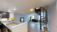 This two-story space offers a thoughtfully designed open-concept layout that seamlessly connects the kitchen and living areas on the main floor. The kitchen features granite countertops with a clean white finish, providing both functionality and a polished aesthetic. Natural light flows generously throughout the main level via multiple windows, illuminating the light hardwood flooring and soft grey walls that lend the space a calm, contemporary feel.

The living area is spacious and inviting, with ample room for seating and furnishings. A standout architectural feature is the sleek black spiral staircase, which serves as both a functional element and a visual centerpiece, connecting the main floor to the upper level with an elegant, space-efficient design. The upper level benefits from its own natural light source, visible through windows that brighten the mezzanine area.

Recessed lighting throughout the main floor ensures consistent illumination, complementing the abundant natural light. The overall design balances modern minimalism with warmth, making it a versatile and functional space suited for both everyday living and entertaining.