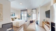 This thoughtfully designed single-floor residence offers a seamless open-concept layout that maximizes both functionality and comfort. Light wood flooring flows throughout the main living areas, creating a warm and cohesive aesthetic anchored by a neutral, contemporary palette.

The kitchen is a standout feature, presented as a well-organized galley-style space with white quartz countertops, subway tile backsplash, and two-toned cabinetry accented by matte black hardware. Stainless steel appliances — including a refrigerator, dishwasher, range, and over-the-range microwave — complete the space. A deep single-basin sink with a matte black faucet adds a refined finishing touch.

The bedroom area is generously proportioned and bathed in natural light from floor-to-ceiling windows fitted with roller shades. Two sliding glass doors provide access to a private balcony with outdoor views. The room comfortably accommodates a king-sized bed and a lounge sofa, with a wall-mounted TV and media console offering additional functionality.

A compact dining nook with a round table and rattan chairs sits adjacent to the sleeping area, offering a defined space for meals. The bathroom features large-format light tile flooring, a floating vanity with ample under-cabinet storage, and a separate walk-in closet, providing excellent storage capacity. Recessed lighting throughout ensures a bright, clean ambiance across all areas of the unit.