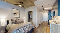 This single-floor urban unit showcases an industrial-modern aesthetic defined by its signature exposed concrete ceiling, which adds raw character throughout the living spaces. Wood-look plank flooring runs continuously across the unit, providing warmth that contrasts beautifully with the concrete overhead.

The bedroom features the exposed concrete ceiling accented by a ceiling fan, track lighting, and an open closet with built-in shelving and hanging rod. The bedroom transitions naturally into the living area, where a large floor-to-ceiling window frames city views and fills the space with natural light. The living area comfortably accommodates a seating arrangement and media setup, with a wall-mounted TV and floating media console.

The galley-style kitchen is well-appointed with two-tone cabinetry — warm wood-grain lower cabinets paired with bold navy upper cabinets — stainless steel appliances, white quartz countertops, a deep undermount sink, and a clean white tile backsplash.

The bathroom features large-format dark tile flooring, a wood-grain vanity with a white countertop, and framed wall art that adds a polished finish. The overall ambiance is contemporary and urban, blending industrial materials with modern design details.