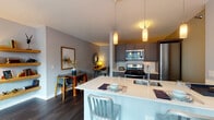 This contemporary single-floor residence showcases a thoughtfully designed open-concept layout that seamlessly integrates the kitchen, dining, and living areas. The kitchen is equipped with stainless steel appliances, including a gas range and built-in microwave, complemented by dark, wood-toned cabinetry and a sleek white quartz island with bar seating—ideal for casual dining or entertaining. Pendant lighting adds warmth above the island and throughout the kitchen.

The living area is a true highlight, anchored by expansive floor-to-ceiling windows that frame impressive urban skyline views, flooding the space with natural light. Dark hardwood flooring runs throughout, providing a rich contrast to the soft gray walls. Floating shelves, a full-length mirror, and carefully curated décor contribute to the modern, urban aesthetic.

The dining nook accommodates intimate gatherings, accented by framed artwork and a decorative sunburst mirror. The bedroom offers a serene retreat with warm ambient lighting, a tufted headboard, and coordinated artwork. The bathroom features a dark wood vanity with a white countertop, a large mirror, and light-toned tile flooring, maintaining the clean, modern sensibility found throughout the entire residence.
