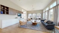 Located at 25550 River Bend Dr in Yorba Linda, CA, this single-floor community space offers a generously sized, thoughtfully designed facility that seamlessly blends comfort and functionality. The layout encompasses two living areas, three exercise rooms, and a connecting hallway, all finished with warm light wood flooring throughout.

The main living lounge is a standout space, characterized by soaring ceilings, abundant natural light streaming through floor-to-ceiling windows, and a modern geometric chandelier that anchors the room. Built-in cabinetry with a combination of white gloss lower units and walnut-toned upper shelving lines one wall, complemented by a wall-mounted television. Contemporary lounge chairs and a wooden octagonal coffee table arranged over a patterned area rug create an inviting gathering area. A secondary living space features a tiled linear fireplace with a mounted television above, flanked by floating wood shelves — adding warmth and a residential character.

The kitchenette area includes a bar-height counter with modern upholstered stools, rich wood cabinetry, stainless steel appliances, and pendant lighting, providing a functional social hub. The fitness center is well-appointed with multiple cardio machines, mirrored walls, and large windows that introduce natural light and outdoor views, creating an energizing environment for exercise.