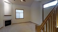 This two-story space is characterized by its vaulted ceilings, which lend an open, airy feel to the upper level. The main living area pictured features soft, neutral-toned carpet flooring complemented by warm wood baseboards and trim that frame the room cohesively. A ceiling fan with a built-in track light fixture is mounted at the peak of the vaulted ceiling, providing both ventilation and overhead lighting. A window dressed with vertical blinds allows natural light to filter into the space, brightening the neutral walls. The staircase, positioned along one side of the room, features natural wood railings and balusters, adding a warm architectural detail that connects the two floors. A transom-style window near the staircase landing introduces additional natural light from above. The overall ambiance of the space is clean and understated, with a simple, functional layout that emphasizes the generous ceiling height and open feel of the upper level. The consistent use of wood tones throughout the trim, railings, and window frames creates a unified, cohesive aesthetic across the space.