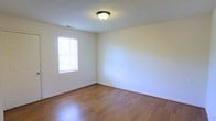 This single-story space presents a straightforward and functional layout with a clean, understated aesthetic. The room visible in the image showcases smooth hardwood-style flooring in a warm honey tone, reflecting light and contributing to an open, airy feel. The walls are finished in a crisp, neutral white, offering a blank canvas adaptable to a variety of uses and décor styles. A textured popcorn ceiling adds a subtle dimensional quality overhead, with a centrally mounted flush light fixture providing consistent ambient illumination. A window fitted with horizontal blinds allows natural light to filter into the room, enhancing the sense of brightness while maintaining privacy. White baseboards frame the perimeter of the room cleanly, complementing the overall neutral palette. The space feels uncluttered and well-maintained, with an electrical outlet visible along one wall. The single-floor layout suggests ease of accessibility and efficient use of space throughout. Overall, the interior conveys a practical, move-in-ready environment suited for residential living.