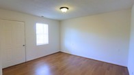 This single-story space presents a straightforward and functional layout with a clean, understated aesthetic. The room visible in the image showcases smooth hardwood-style flooring in a warm honey tone, reflecting light and contributing to an open, airy feel. The walls are finished in a crisp, neutral white, offering a blank canvas adaptable to a variety of uses and décor styles. A textured popcorn ceiling adds a subtle dimensional quality overhead, with a centrally mounted flush light fixture providing consistent ambient illumination. A window fitted with horizontal blinds allows natural light to filter into the room, enhancing the sense of brightness while maintaining privacy. White baseboards frame the perimeter of the room cleanly, complementing the overall neutral palette. The space feels uncluttered and well-maintained, with an electrical outlet visible along one wall. The single-floor layout suggests ease of accessibility and efficient use of space throughout. Overall, the interior conveys a practical, move-in-ready environment suited for residential living.