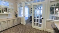 This single-floor space exudes a polished, traditional aesthetic that balances professional functionality with refined design. The standout architectural feature is a set of classic French doors with multi-pane glass panels and a matching transom window above, allowing light to flow freely between areas while maintaining a sense of defined space. Tall windows dressed with slatted blinds and flowing curtains flood the interior with natural light, enhancing the warm, neutral color palette throughout. The office area beyond the French doors is furnished with ergonomic seating and a working desk, making it well-suited for private meetings or focused work. Patterned carpeting in muted tones runs throughout, adding texture and a sense of warmth underfoot. Built-in cabinetry with a distressed, painted finish provides practical storage and lends a charming, cottage-inspired detail to the space. A decorative chandelier visible through the glass doors adds an elegant finishing touch, while a floral arrangement on the cabinetry introduces a subtle natural element. Overall, the space conveys a calm, professional ambiance with thoughtful design details that feel both functional and inviting.
