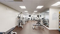 Located at 2249 NJ-37 E in Toms River, New Jersey, this fitness center offers a well-equipped, single-floor workout facility designed to support a comprehensive exercise routine. The space features a drop-panel ceiling with recessed fluorescent lighting that ensures even, bright illumination throughout. Durable, speckled rubber flooring covers the entire workout area, providing both cushioning and traction during exercise.

The main workout area is generously sized and divided into distinct functional zones. The cardio section is lined with a row of True treadmills and elliptical machines, each equipped with individual display consoles and positioned beneath wall-mounted flat-screen televisions. A dedicated strength training zone houses a Hoist multi-station cable machine, a selectorized resistance unit, and additional strength equipment. The free weights area features a full rack of dumbbells paired with adjustable benches, while a medicine ball rack and stability balls offer options for functional training.

Full-length mirrors along one wall visually expand the space and assist with form during workouts. A reception or check-in counter is visible near the entrance, and the facility includes an ADA-accessible restroom with grab bars, a wall-mounted sink, and classic hexagonal tile flooring. A secondary entrance with a door and window provides additional access and natural light. Overall, the space presents a clean, organized, and functional environment suitable for a wide range of fitness activities.