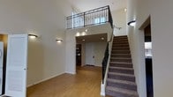 This space makes an immediate impression with its dramatic double-height ceilings that flood the main living area with a sense of openness and volume. The entry level showcases warm, tile flooring in a classic brick-like pattern that extends throughout the ground floor, complementing the neutral, light-toned walls.

A carpeted staircase with a sleek black wrought-iron railing leads to an upper-level loft or mezzanine, which overlooks the main floor through an ornate, scrollwork iron balustrade — a distinctive architectural detail that adds elegance to the space. The upper level benefits from natural light streaming in through a window, brightening the full height of the interior.

Recessed lighting and wall-mounted sconces provide layered illumination throughout. The lower level transitions into a secondary area featuring additional recessed lighting, built-in appliances visible in the background, and a white entry door, suggesting a functional kitchen or utility zone nearby.

Overall, the space balances contemporary practicality with refined architectural details, creating an inviting and visually dynamic interior.