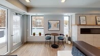 This well-appointed single-floor amenity lounge offers a thoughtfully designed space that blends entertainment, relaxation, and functionality. The open layout is anchored by warm wood-look flooring that runs throughout, creating a cohesive and inviting atmosphere.

A standout feature is the bold stone-tile accent wall, which serves as the backdrop for a large wall-mounted flat-screen television — making it an ideal focal point for social gatherings. Adjacent to the entertainment area, a shuffleboard table adds a recreational element, while high-top bistro tables with teal upholstered stools provide casual seating options.

Large windows along one wall flood the space with natural light and offer views of the surrounding urban streetscape. Potted greenery on the windowsills adds a touch of warmth and life to the contemporary aesthetic. Contemporary artwork adorns the walls, contributing to the space's modern character.

The kitchenette is fully equipped with rich dark-wood cabinetry, dark stone countertops, a white subway tile backsplash, stainless steel appliances — including a refrigerator, microwave, and dishwasher — and a sink, making it convenient for hosting events. A horizontal shiplap accent wall adds architectural texture. The lounge also features a comfortable seating arrangement with sofas and ottomans, perfect for relaxed conversation.