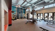 This fitness center offers a spacious, single-floor layout designed to accommodate a comprehensive range of workout routines. The space is defined by its striking soaring ceilings with exposed silver ductwork, lending an industrial-modern aesthetic that feels open and energizing. Distinctive circular pendant lights in deep red hang overhead, complementing the bold teal and red accent walls that divide the space into visually dynamic zones.

The cardio area features a row of True treadmills positioned near floor-to-ceiling glass doors, which flood the space with natural light and provide views to the outdoors. A wall-mounted flat-screen TV accompanies this section for entertainment during workouts. The strength training area is anchored by a multi-functional cable machine and includes adjustable benches, a StairMaster, and a dedicated dumbbell rack with a full weight selection. A functional training zone offers TRX suspension straps, kettlebells, medicine balls, and a heavy bag stored on a built-in wall-mounted rack. A built-in seating nook with cushioned panels adds a thoughtful lounge element. Rubber flooring throughout ensures durability and safety. Large windows along the perimeter bring in abundant natural light, enhancing the overall inviting and motivating atmosphere of this well-appointed fitness facility.
