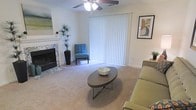 This single-floor space showcases a thoughtfully designed living area that balances comfort and style. The room centers around a standout marble-surround fireplace, which serves as an elegant focal point along one wall, adding a classic architectural detail to the space. Soft, neutral-toned wall-to-wall carpeting runs throughout, contributing to the warm and inviting ambiance underfoot.

A ceiling fan with an integrated light fixture is mounted overhead, providing both practical air circulation and ambient lighting. Large vertical blinds span a generous window or sliding door along one wall, filtering in abundant natural light while offering privacy and light control. The brightness these blinds allow gives the room an open, airy feel.

The living area is well-proportioned, comfortably accommodating a sofa, an accent chair in a bold teal upholstery, and an oval coffee table with a sleek metal base. Decorative touches, including framed wall art and a tall potted plant near the fireplace, add personality and a sense of life to the space. The overall aesthetic is clean, approachable, and move-in ready.