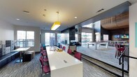This single-floor amenity lounge is designed to accommodate both social gatherings and casual recreation. The space is organized into distinct functional zones that flow naturally into one another. A long, white communal table anchored by a bold yellow pendant light serves as a central gathering point, surrounded by deep red upholstered chairs that add a pop of color against the neutral grey carpeting and light-toned walls.

Adjacent to the seating area, a pool table with a striking red felt surface provides a recreational focal point. The space benefits from generous natural light streaming through large floor-to-ceiling windows and glass doors along multiple walls, offering views of an outdoor patio area furnished with seating. This connection between indoor and outdoor spaces enhances the overall openness of the lounge.

The interior design balances modern and industrial elements, featuring dark accent wall panels, recessed lighting, and visible HVAC components that contribute to a contemporary aesthetic. A lounge seating area with a large-screen television occupies one corner, rounding out the entertainment options. The overall ambiance is energetic yet functional, making it well-suited for both group activities and relaxed socializing.