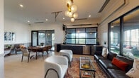 Located at 1512 Bull St in the heart of Savannah, Georgia, this single-floor amenity lounge delivers a refined blend of contemporary design and Southern charm. The open-plan layout is anchored by a striking living area featuring a dark leather sofa, plush boucle accent chairs, and a glass coffee table, all centered beneath a sculptural gold-and-globe chandelier that serves as a bold focal point overhead.

A defining architectural feature is the industrial-style glass garage door that spans one wall, opening onto a furnished outdoor terrace. This seamless indoor-outdoor connection floods the interior with natural light and offers tree-lined views of the surrounding Savannah landscape. Large sliding glass doors further enhance this connection to the terrace, which is outfitted with additional seating and greenery.

Along the interior wall sits a sleek wet bar with dark cabinetry, light countertops, gold hardware, a built-in sink, and a stainless steel beverage refrigerator — ideal for entertaining. Elegant wainscoting and crown molding line the walls throughout, adding a classic architectural detail that complements the modern furnishings. A dedicated dining area with a walnut table and mixed seating, along with a quiet sitting nook adorned with framed artwork, round out this well-appointed and versatile community space.