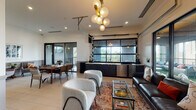 Located at 1512 Bull St in the heart of Savannah, Georgia, this single-floor amenity lounge delivers a refined blend of contemporary design and Southern charm. The open-plan layout is anchored by a striking living area featuring a dark leather sofa, plush boucle accent chairs, and a glass coffee table, all centered beneath a sculptural gold-and-globe chandelier that serves as a bold focal point overhead.

A defining architectural feature is the industrial-style glass garage door that spans one wall, opening onto a furnished outdoor terrace. This seamless indoor-outdoor connection floods the interior with natural light and offers tree-lined views of the surrounding Savannah landscape. Large sliding glass doors further enhance this connection to the terrace, which is outfitted with additional seating and greenery.

Along the interior wall sits a sleek wet bar with dark cabinetry, light countertops, gold hardware, a built-in sink, and a stainless steel beverage refrigerator — ideal for entertaining. Elegant wainscoting and crown molding line the walls throughout, adding a classic architectural detail that complements the modern furnishings. A dedicated dining area with a walnut table and mixed seating, along with a quiet sitting nook adorned with framed artwork, round out this well-appointed and versatile community space.