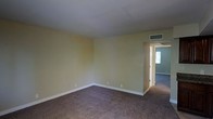 This single-floor space presents a straightforward and functional layout. The main room is finished with wall-to-wall carpet in a muted brown tone, providing a comfortable underfoot feel throughout. The walls are painted in a soft yellow-green hue, complemented by crisp white baseboards that run along the perimeter, lending the space a clean and well-maintained appearance. A ceiling-mounted HVAC vent ensures climate control, while the smooth white ceiling above contributes to a bright, open atmosphere. An open doorway leads directly into an adjoining room, which continues the same carpeted flooring and neutral color palette, suggesting a cohesive design throughout the space. The connecting room benefits from natural light filtering in through a visible window, brightening the interior. The overall layout is open and unobstructed, making the space highly adaptable for a variety of uses. The simple, no-frills design provides a neutral canvas that can be easily personalized. The single-floor configuration ensures convenient, step-free accessibility throughout the entire space.