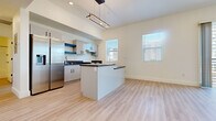 This contemporary single-floor residence showcases a clean, modern design with light-toned luxury vinyl plank flooring extending throughout every room, creating a cohesive and airy atmosphere. The open-plan kitchen and living area serves as the heart of the space, anchored by a peninsula with a dark countertop that doubles as a breakfast bar. The kitchen is well-appointed with white flat-front cabinetry, open floating shelves, stainless steel appliances including a refrigerator and range, and a sleek undermount sink. A striking geometric pendant light fixture hangs above, adding a modern accent. Soaring ceilings in the main living area amplify the sense of openness. Multiple windows fitted with horizontal blinds invite abundant natural light throughout the unit. The bedrooms are generously proportioned, each featuring large windows that frame views of the surrounding urban cityscape, with one room offering access to a private balcony through full-height sliding glass doors. The bathroom is finished with a clean, all-white walk-in shower surround, complemented by chrome fixtures and a dark-topped vanity. Recessed lighting and matte black door hardware provide refined finishing touches that carry the modern aesthetic consistently through the entire space.