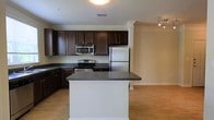 Located at 5806 Ogeechee Rd in Savannah, Georgia, this single-floor space offers a well-designed, open-concept layout that blends functionality with modern style. The kitchen serves as a central highlight, featuring rich dark wood cabinetry that extends to the ceiling, providing generous storage throughout. A large center island with a dark granite-style countertop offers additional prep space and serves as a natural gathering point. Stainless steel appliances — including a refrigerator, electric range, and built-in microwave — lend a sleek, contemporary feel to the cooking area. The kitchen flows seamlessly into the adjacent living area, creating an open and connected environment ideal for entertaining. Crown molding frames the ceilings throughout, adding a refined architectural touch, while the warm hardwood-style flooring extends across the main living spaces, bringing cohesion to the interior. A track lighting fixture in the living area provides adjustable illumination, complementing the overall bright and airy atmosphere. The textured ceiling and neutral wall tones create a clean, versatile backdrop throughout the space.