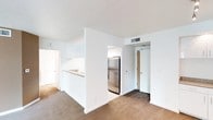 This single-floor space is designed with a functional, open layout that promotes a sense of connectivity between living areas. The main living area features neutral carpet flooring and crisp white walls, creating a clean and understated aesthetic. A half-wall partition separates the living space from the kitchen, which is accessible via a pass-through counter ideal for casual dining or food preparation. The kitchen is equipped with stainless steel appliances, including a full-sized refrigerator, and features white cabinetry along with a sink positioned at the pass-through. The flooring transitions from carpet in the living area to a hard surface in the kitchen zone, providing clear delineation between spaces. HVAC vents are integrated into the ceiling, ensuring climate control throughout. A doorway visible from the living area leads to additional rooms, while a secondary door with a warm wood finish is accessible from the kitchen area, likely serving as a utility or storage entry. The overall design prioritizes practicality with a neutral color palette, white trim, and straightforward finishes that offer a versatile foundation for personalization.