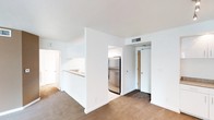 This single-floor space is designed with a functional, open layout that promotes a sense of connectivity between living areas. The main living area features neutral carpet flooring and crisp white walls, creating a clean and understated aesthetic. A half-wall partition separates the living space from the kitchen, which is accessible via a pass-through counter ideal for casual dining or food preparation. The kitchen is equipped with stainless steel appliances, including a full-sized refrigerator, and features white cabinetry along with a sink positioned at the pass-through. The flooring transitions from carpet in the living area to a hard surface in the kitchen zone, providing clear delineation between spaces. HVAC vents are integrated into the ceiling, ensuring climate control throughout. A doorway visible from the living area leads to additional rooms, while a secondary door with a warm wood finish is accessible from the kitchen area, likely serving as a utility or storage entry. The overall design prioritizes practicality with a neutral color palette, white trim, and straightforward finishes that offer a versatile foundation for personalization.