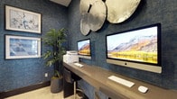 This single-floor business center is thoughtfully designed to provide a productive and visually engaging work environment. The space is defined by its striking deep-blue textured wallcovering, which lends a sophisticated and cohesive aesthetic throughout. Multiple workstations are arranged along sleek, dark-toned desks, each equipped with iMac computers and supported by ergonomic task chairs in warm tan upholstery, offering both comfort and functionality. A printer station is conveniently available for shared use.

Decorative elements elevate the space beyond a typical office setting. A cluster of metallic disc wall sculptures adds an artistic focal point above one of the workstations, while a curated gallery of four framed artworks in blue and white tones adorns another wall, reinforcing the cohesive color palette. Tall potted bamboo plants introduce a natural, refreshing contrast to the polished interior finishes.

Cabinetry with neutral countertops provides built-in storage and additional workspace. A pendant light fixture overhead and access to an adjacent room with visible natural light through glass doors contribute to an open and well-lit atmosphere. The light-toned carpeting grounds the room with a clean, professional finish, making this business center a refined and fully equipped space for productivity.