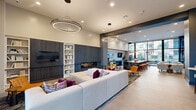 Located at 1345 14th St in Plano, Texas, this sophisticated single-floor community living space offers a generously sized, open-plan layout designed for both socializing and relaxation. The space is anchored by a sleek, contemporary aesthetic, featuring large-format tile flooring, exposed structural beams with dark accents, and soaring ceilings that contribute to an expansive, airy atmosphere throughout.

The living area is thoughtfully divided into distinct zones. A plush sectional sofa with vibrant accent pillows anchors the main lounge, positioned beside a slatted accent wall housing a flat-screen TV and a linear fireplace. An adjacent built-in bookshelf adds a library-inspired reading nook with comfortable curved chairs nearby. The game zone features a full-size billiards table and a shuffleboard, making it a standout entertainment hub.

A dramatic black marble island serves as the centerpiece of the social dining area, surrounded by stylish seating and pendant lighting. Floor-to-ceiling windows along multiple walls flood the space with natural light and offer expansive views of the surrounding outdoor area and cityscape.

The entrance foyer welcomes visitors with curated abstract artwork, a cozy built-in bench, and glass-panel doors. Eclectic furnishings, bold artwork displays, and designer lighting fixtures are woven throughout, creating a cohesive, upscale ambiance ideal for community gatherings.