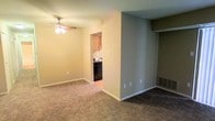 This single-floor residential space offers a practical and functional layout designed for comfortable everyday living. The main living area is finished with wall-to-wall carpeting that adds warmth underfoot, while a ceiling fan with integrated lighting provides both air circulation and ambient illumination. Neutral-toned walls throughout create a clean, versatile backdrop that adapts easily to a variety of décor styles. A partially open partition wall separates the living area from the kitchen, maintaining a sense of connectedness between the two spaces while providing a degree of visual distinction. The kitchen features wood-toned upper cabinetry and is equipped with black appliances, including a dishwasher, lending a classic contrast to the lighter cabinetry. The kitchen flooring transitions to a smooth hard surface, differentiating it from the carpeted living zone. A recessed shelving or closet area is visible along one wall of the living space, offering convenient built-in storage. Floor-level air vents are integrated throughout, supporting the home's climate control system. The overall layout is straightforward and efficient, making it an approachable and easy-to-maintain living environment.