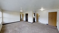 This single-floor unit offers a straightforward and functional layout designed for comfortable everyday living. The main living area is spacious and open, featuring wall-to-wall carpeting in a neutral tone that adds warmth underfoot, complemented by clean white walls and a textured ceiling. Flush-mounted ceiling lights provide steady illumination throughout the space.

A central structural column defines the transition between the living area and the kitchen zone, creating a natural sense of separation without fully closing off the spaces. The kitchen is equipped with dark cabinetry, a stainless steel refrigerator, and a sink, offering practical functionality for meal preparation. Adjacent to the kitchen, a washer unit is visible, indicating in-unit laundry convenience.

Doorways visible from the main living area lead to additional rooms, suggesting a connected layout that likely includes a bedroom and bathroom. One doorway reveals a glimpse of a bathroom with light-colored fixtures and cabinetry, while another opens toward what appears to be a bedroom or hallway. The overall ambiance is clean, neutral, and move-in ready, with a simple aesthetic that provides a versatile canvas for personalization. Baseboard heaters along the walls offer a dedicated heating source throughout the unit.