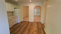 This single-floor residence offers a welcoming and functional layout with thoughtful architectural details throughout. The kitchen is outfitted with white cabinetry, ample counter space, and a full-size refrigerator, providing a clean and practical cooking environment. Rich, wide-plank hardwood flooring runs throughout the main area, adding warmth and character to the space.

A distinctive arched entryway serves as a centerpiece of the interior, gracefully connecting the kitchen and main living areas to an adjoining room. This architectural detail lends a classic charm to the overall design. The adjoining room features plush carpeting and is bathed in natural light through a double-pane window that frames views of the surrounding greenery, creating a serene and inviting atmosphere.

A square doorway adjacent to the arch leads to an additional carpeted room, offering flexible use as a bedroom, office, or secondary living space. The neutral wall tones throughout complement both the hardwood and carpeted floors, creating a cohesive and understated aesthetic. Recessed lighting in the kitchen area ensures the space is well-lit. Overall, this home presents a blend of classic design elements and practical amenities in a comfortable, single-level layout.