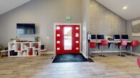 Located at 9600 E Girard Ave in Denver, Colorado, this single-floor amenity and leasing space serves the Birge & Held–managed Grammercy Fitness community. The interior is defined by soaring vaulted ceilings that lend an open, airy feel throughout the main area, which is finished with wood-look tile flooring in a warm, neutral tone. The color palette is crisp grey and white, accented throughout by bold red furnishings that give the space a modern, energetic character.

Upon entry through the striking red double doors, residents and visitors are welcomed by a branded Birge & Held entryway mat and a resident services station equipped with computer terminals and high-top red seating. The central gathering area features a stylish island with a patterned tile surround and bar-height red stools, alongside a full kitchenette with white cabinetry, a sink, and small appliances — ideal for resident use and community events. A wall-mounted flat-screen TV and pendant lighting further enhance the lounge atmosphere.

A dedicated private office provides a quiet consultation area, furnished with a wood-and-metal desk, red guest chairs, and large windows that invite natural light. An adjacent lounge area offers comfortable seating, a wall-mounted TV, and direct access to the outdoors. Glass doors connect the main space to the Grammercy Fitness center, keeping wellness central to the experience.
