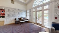 Located at 4122 Avalon Dr in Peabody, MA, this single-floor clubhouse space is a well-appointed gathering destination designed for both relaxation and entertainment. The living room is anchored by soaring ceilings with coffered detailing, a classic fireplace framed in white millwork, and an elegant arched entryway. A sweeping arched transom window floods the space with natural light, while French doors open directly to an outdoor area, seamlessly connecting interior and exterior. Sophisticated cloud-motif wallpaper lines the walls alongside crisp wainscoting, and a bold patterned carpet grounds the room. A red billiards table serves as a striking centerpiece, complemented by a wall-mounted television and curated seating. The dining room transitions into a vibrant game room, distinguished by deep charcoal walls beneath a dramatic orange ceiling. A round poker table sits at the heart of the room, surrounded by sleek black leather chairs. Wall-mounted Scrabble and tic-tac-toe boards add a playful, interactive dimension to the space. Accent tables in red add pops of color throughout, while patterned carpet and a statement pendant light complete the bold, curated aesthetic of this dynamic and inviting space.