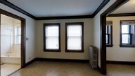 This single-floor space showcases a blend of classic architectural character and functional design. The interior is defined by striking dark wood trim that frames the windows, doorways, and baseboards, creating a bold contrast against the light cream-colored walls and bright white ceiling. Crown molding runs along the ceiling perimeter, adding a refined, traditional touch to the overall aesthetic.

Natural light enters generously through two double-hung windows fitted with horizontal blinds, illuminating the speckled terrazzo-style flooring that spans the room. The flooring features a neutral, multi-toned pattern that complements the room's classic color palette and adds visual texture underfoot.

A vintage cast-iron radiator is positioned along one wall, serving as both a functional heating element and a nod to the building's historic character. The radiator's ornate design is consistent with the traditional architectural language found throughout the space.

The room presents a clean, open layout with no furnishings, making it easy to assess its spatial potential. The combination of natural light, heritage details, and neutral tones creates an atmosphere that is both welcoming and versatile, well-suited for a variety of uses.