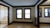 This single-floor space showcases a blend of classic architectural character and functional design. The interior is defined by striking dark wood trim that frames the windows, doorways, and baseboards, creating a bold contrast against the light cream-colored walls and bright white ceiling. Crown molding runs along the ceiling perimeter, adding a refined, traditional touch to the overall aesthetic.

Natural light enters generously through two double-hung windows fitted with horizontal blinds, illuminating the speckled terrazzo-style flooring that spans the room. The flooring features a neutral, multi-toned pattern that complements the room's classic color palette and adds visual texture underfoot.

A vintage cast-iron radiator is positioned along one wall, serving as both a functional heating element and a nod to the building's historic character. The radiator's ornate design is consistent with the traditional architectural language found throughout the space.

The room presents a clean, open layout with no furnishings, making it easy to assess its spatial potential. The combination of natural light, heritage details, and neutral tones creates an atmosphere that is both welcoming and versatile, well-suited for a variety of uses.