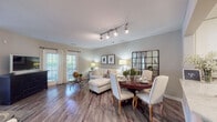 This single-floor residence offers a thoughtfully designed open-concept layout that seamlessly connects the living, dining, and kitchen areas. Rich hardwood-style flooring flows throughout the main living spaces, complemented by soft neutral walls that create a warm, inviting ambiance. The combined living and dining area is anchored by a stylish round dining table paired with upholstered chairs, while a comfortable sectional sofa and track lighting overhead add both function and character. French doors in the living area invite abundant natural light and provide access to the outdoors. The kitchen is well-equipped with white shaker cabinetry, marble-look quartz countertops, a mosaic tile backsplash, and stainless steel appliances including a gas range, microwave, and refrigerator. The primary bedroom is generously sized with plush carpet, large windows dressed in sheer curtains that fill the room with natural light, and ample space for a full dresser suite. A secondary bedroom offers a cozy retreat with soft carpet and serene neutral tones. The bathroom features a clean, modern aesthetic with white cabinetry, a large vanity mirror, and a shower-tub combination. Throughout the residence, consistent design choices and quality finishes create a cohesive and comfortable living environment.