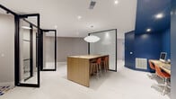 This thoughtfully designed office space occupies a single floor and offers a contemporary, professional environment that balances open collaboration with private workspaces. The layout centers around a sleek natural wood communal workstation topped with built-in power access and paired with vibrant terracotta-hued bar stools, anchored beneath a statement globe pendant light. A bold navy blue accent wall serves as a striking design focal point, housing a built-in wood desk with dual workstations, open shelving, and integrated cabinetry that includes printer storage — providing a fully functional business center within the space. Black-framed glass partition walls and pivot doors define private meeting or focus pods with warm burnt-orange interior walls, offering visual separation without sacrificing openness or light. Recessed ceiling lighting throughout ensures a bright, well-lit atmosphere, complemented by clean white tile flooring that enhances the sense of spaciousness. The design palette — combining crisp whites, deep navy, warm wood tones, and terracotta — creates a cohesive, modern aesthetic that is both professional and visually engaging, making this office well-suited for dynamic, collaborative work environments.