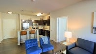 This single-floor residence showcases an open-concept layout that integrates the living and kitchen areas into one cohesive, functional space. The kitchen is outfitted with dark espresso cabinetry, stainless steel appliances including a refrigerator, microwave, and dishwasher, and is anchored by a peninsula-style breakfast bar with seating for multiple guests. Pendant lighting hangs above the counter, adding a warm, inviting glow to the space. The kitchen also features a dedicated sink along the back wall, ample counter space, and open shelving above the upper cabinets for decorative storage. Flowing seamlessly from the kitchen, the living area is finished with hardwood-style flooring that runs throughout, contributing to a clean and modern aesthetic. The living space is well-proportioned, accommodating seating arrangements comfortably. Neutral-toned walls provide a versatile backdrop, while soft ambient lighting from a floor lamp enhances the warmth of the room. A standard interior door near the kitchen provides access to an adjacent room, adding to the functional layout of this well-designed single-level space.
